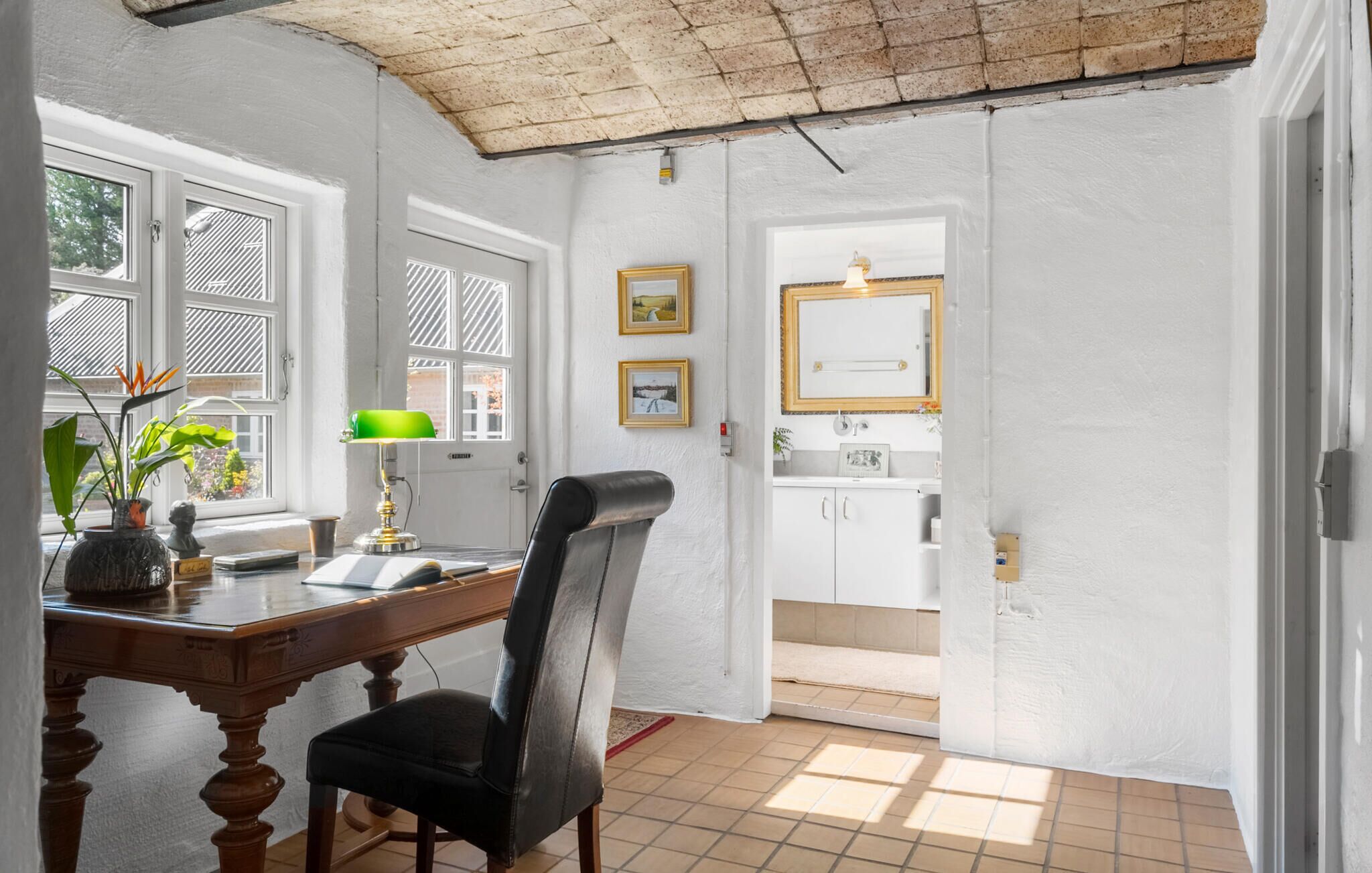 Una habitación con un escritorio de madera, una silla de cuero negro, una lámpara de escritorio verde y una ventana con vista a árboles.