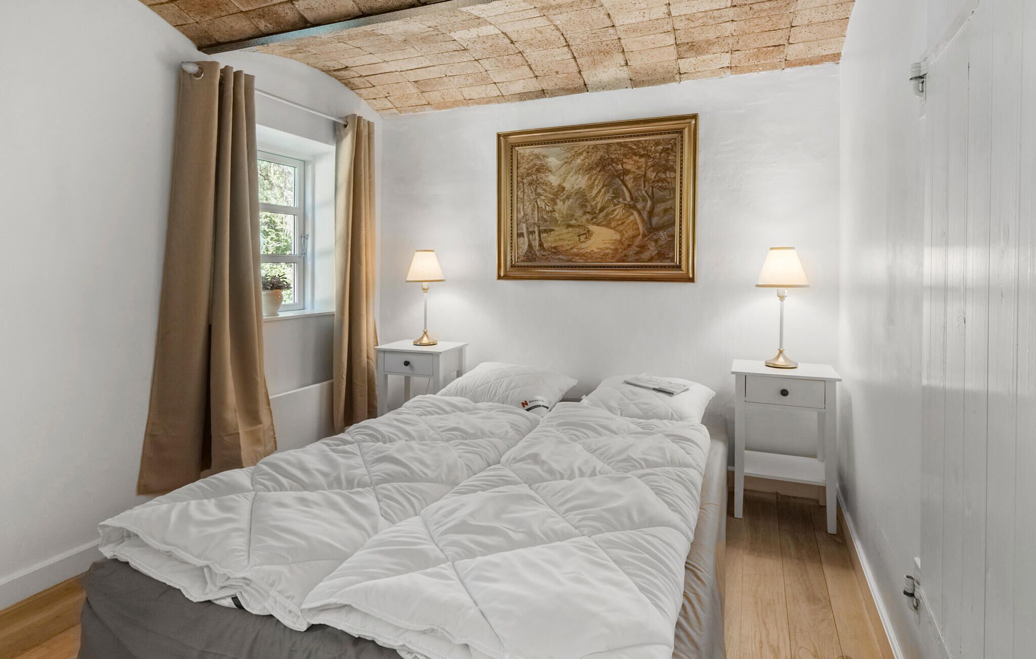 Een slaapkamer met een bed, twee nachtkastjes met lampen, een raam met gordijnen, en een lijst met een schilderij aan de muur.