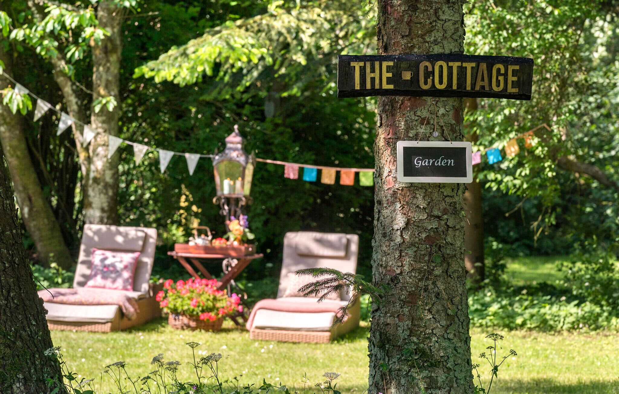 A sign reading "THE COTTAGE" and "Garden" in a lush, green outdoor setting with a picnic table and lounge chairs.