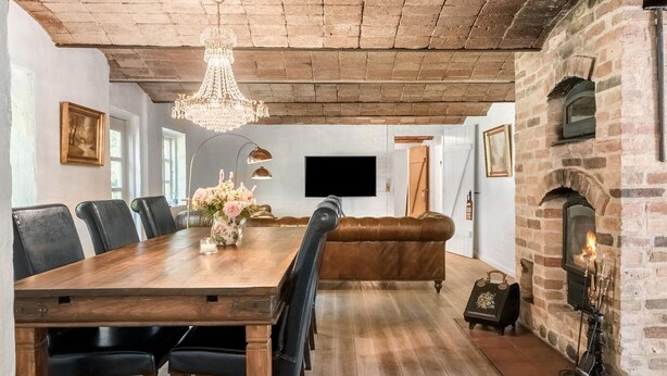A dining area with a wooden table, leather chairs, a chandelier, a fireplace, and a television mounted on the wall.