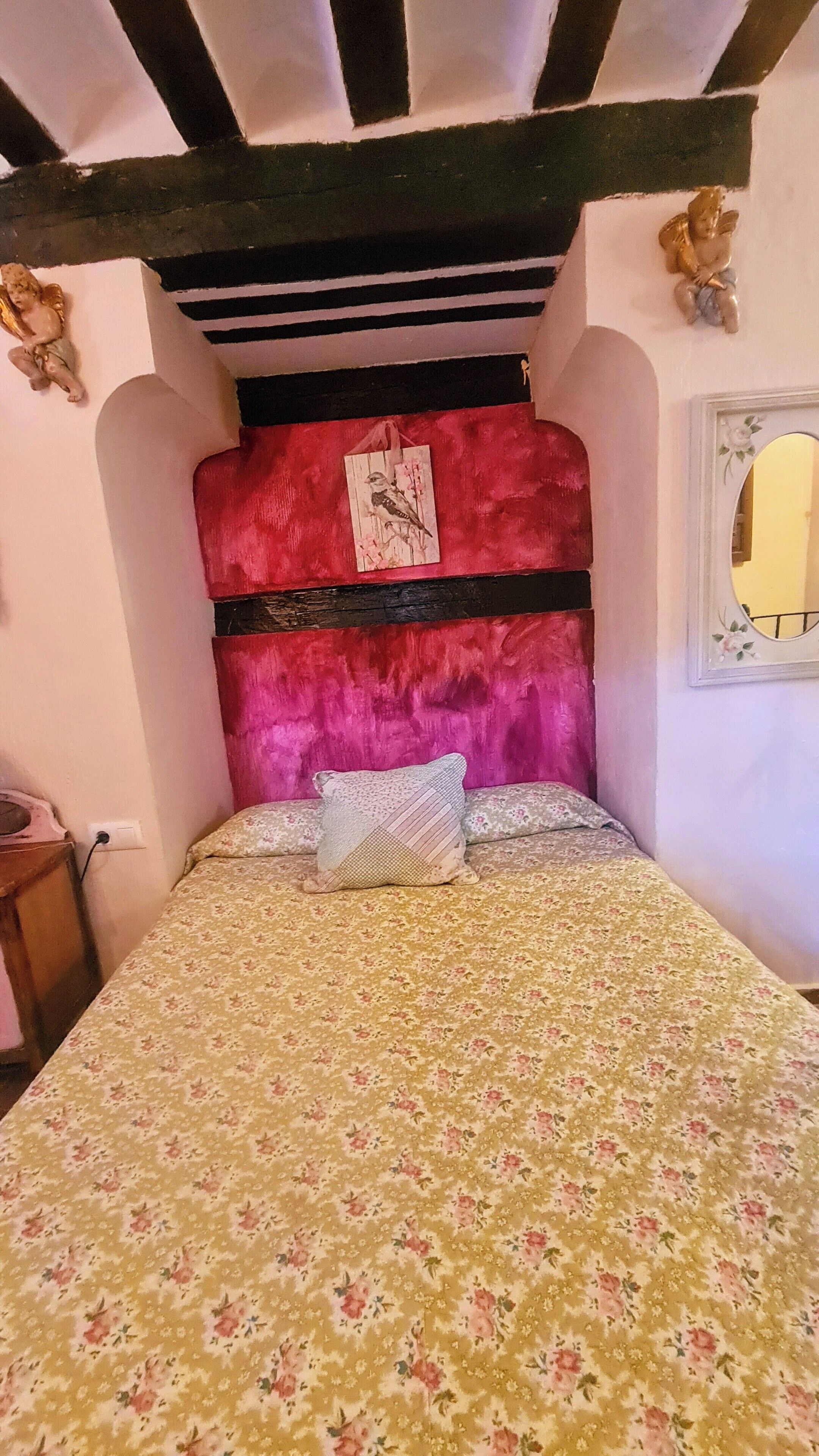 Un dormitorio con colcha estampada, cabecera tapizada en rojo y un pequeño ángel decorativo.