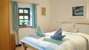 A bedroom with a bed, a window with curtains, a radiator, a bedside table, and a wall with a framed picture. - Daisy Cottage Berrynarbor near Broadsands beach (Berrynarbor)