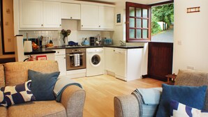 A compact kitchen with white cabinets, a washing machine, and a dishwasher. There are two sofas with patterned cushions in the living area. - Daisy Cottage Berrynarbor near Broadsands beach (Berrynarbor)