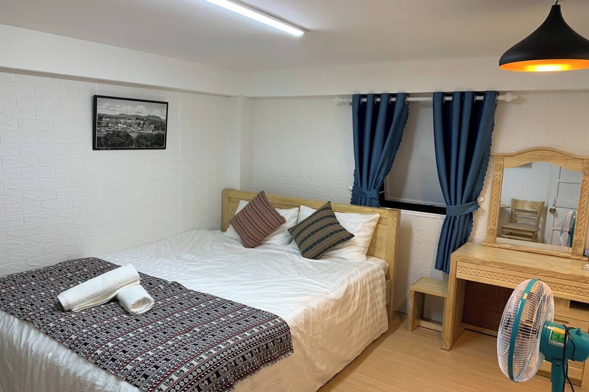 Une chambre d’hôtel avec un lit, une table de chevet, un miroir, un ventilateur et une fenêtre avec des rideaux.