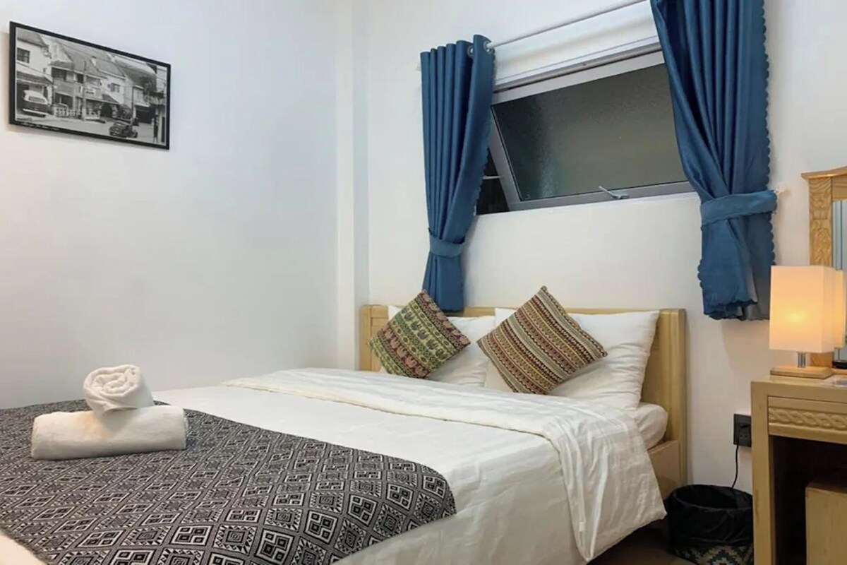 Habitación de hotel con cama, ventana con cortinas, mesita de noche con lámpara y un cuadro colgado en la pared.