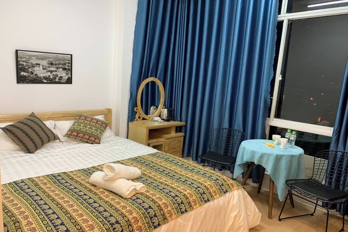 Une chambre d’hôtel comprenant un lit, un bureau avec un miroir, une chaise, une petite table recouverte d’une nappe et une fenêtre avec des rideaux.