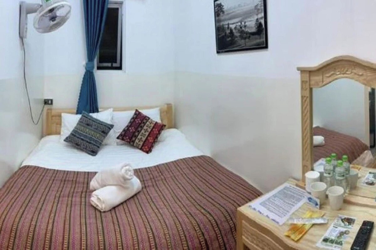 A hotel room with a bed, a bedside table with toiletries, a wall-mounted fan, and a window with curtains.