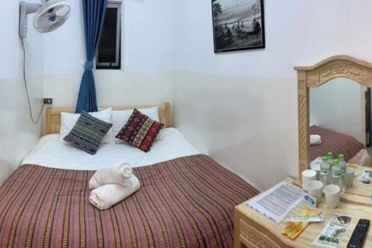 Une chambre d’hôtel comprenant un lit, une table de chevet avec des articles de toilette, un ventilateur fixé au mur et une fenêtre avec des rideaux.