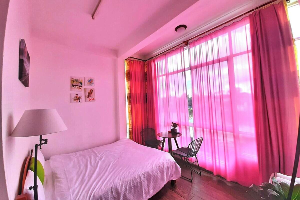 A room with a bed, a table, two chairs, and a window with pink curtains.