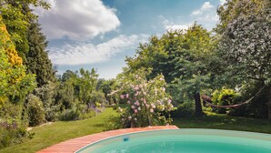 Ein Schwimmbecken mit Holzterrasse, umgeben von üppiger Vegetation und blühenden Bäumen.