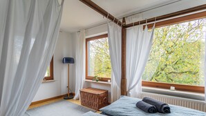 Ein Schlafzimmer mit zwei Betten, einem Korbwäscheschrank, einer Stehlampe und Blick auf Bäume durch das Fenster.