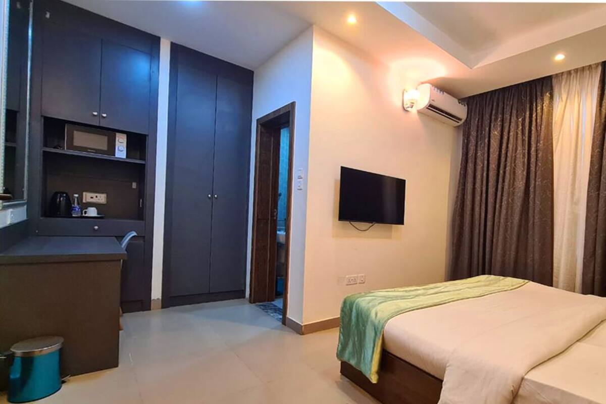 A modern hotel room with a bed, a TV mounted on the wall, a kitchenette, and a large window with curtains.