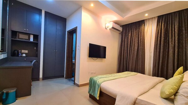 Ein modernes Hotelzimmer mit Bett, Wand-Fernseher, einer Küchenzeile und einem großen Fenster mit Vorhängen.