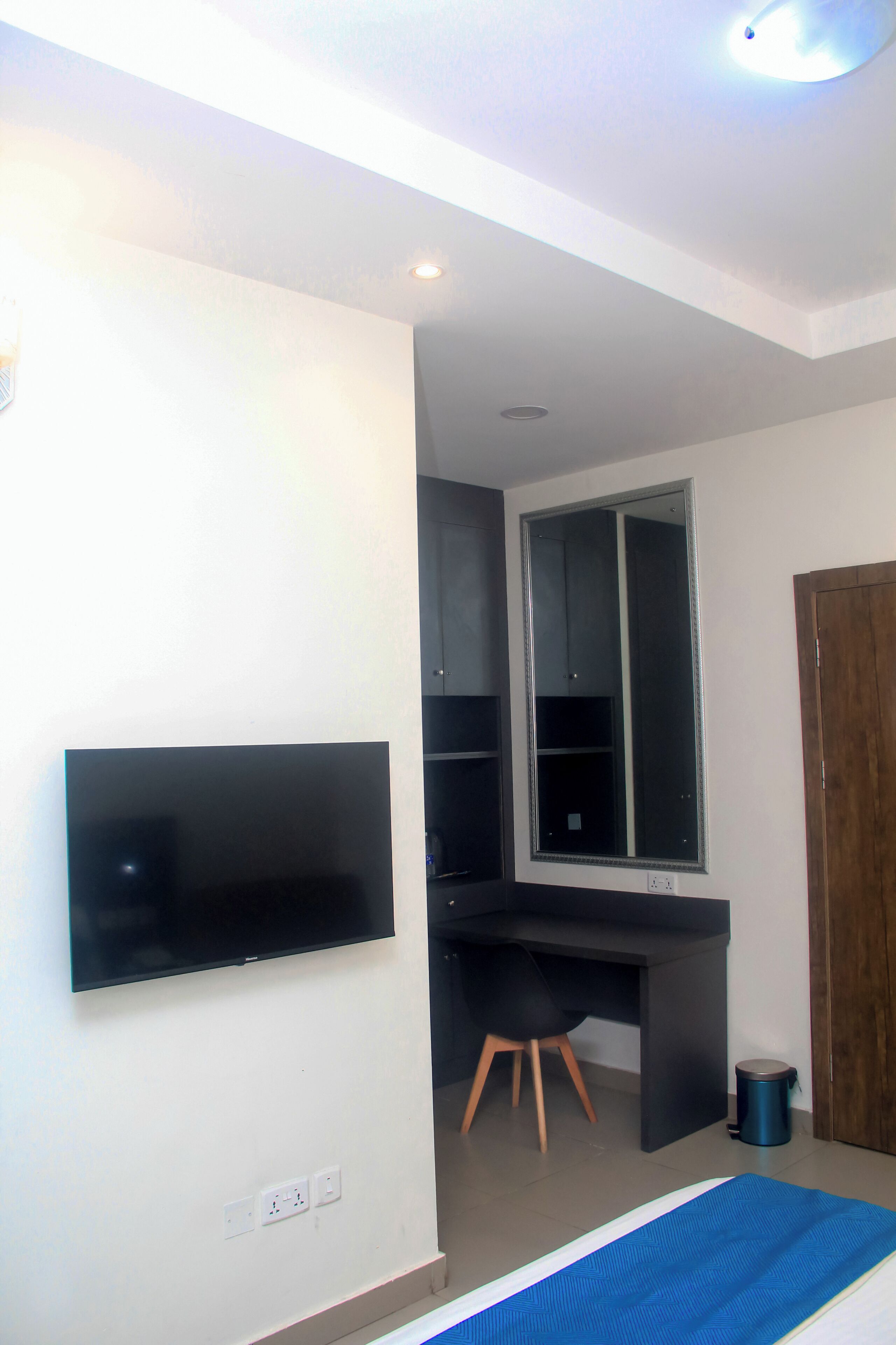 A room with a flat-screen TV mounted on the wall, a desk with a chair, a large window, and a door.