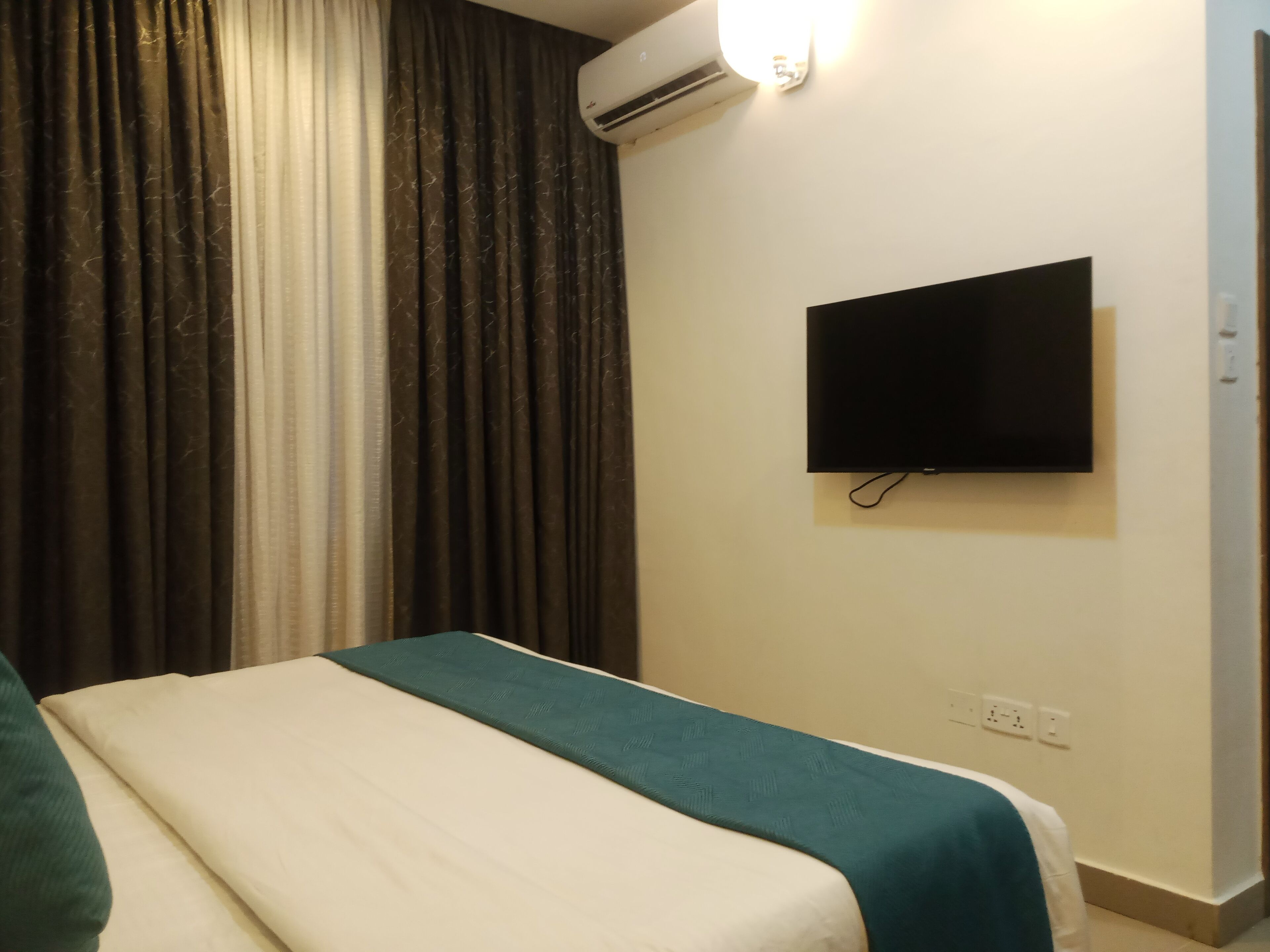 A hotel room with a bed, a TV mounted on the wall, and an air conditioning unit.