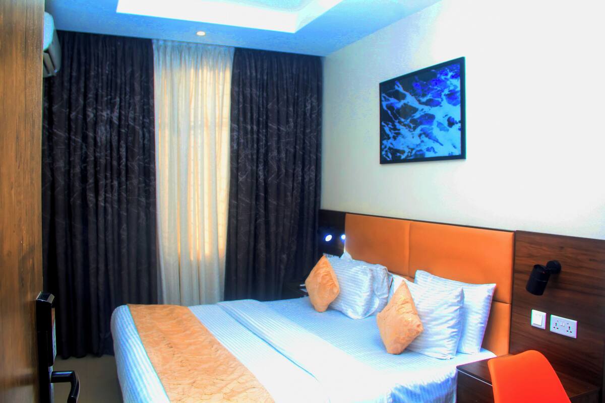 A hotel room with a bed, curtains, a wall-mounted artwork, and a nightstand.