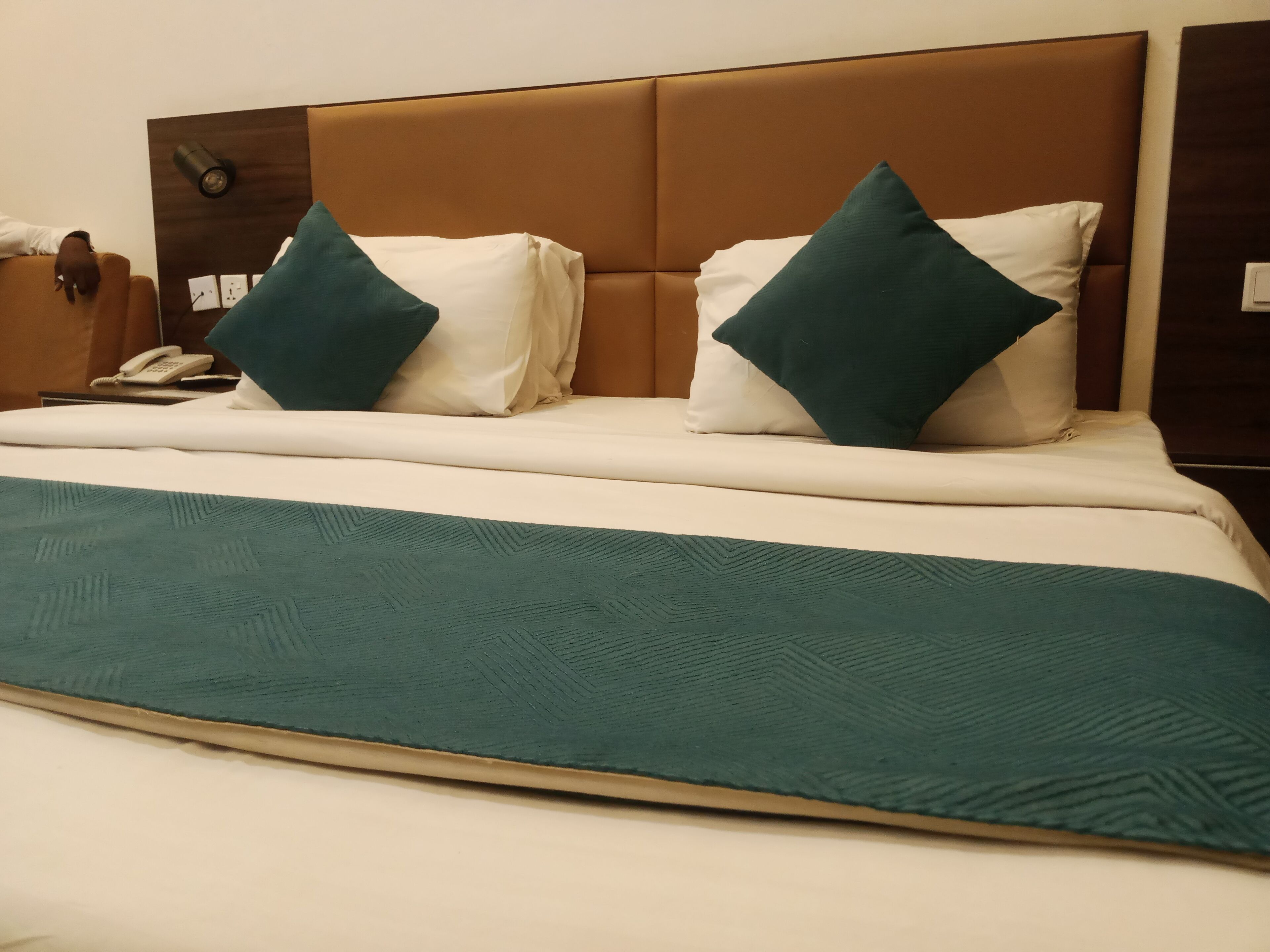 A neatly made bed with a teal blanket and two pillows, a wooden headboard, a bedside phone, and a wall clock.
