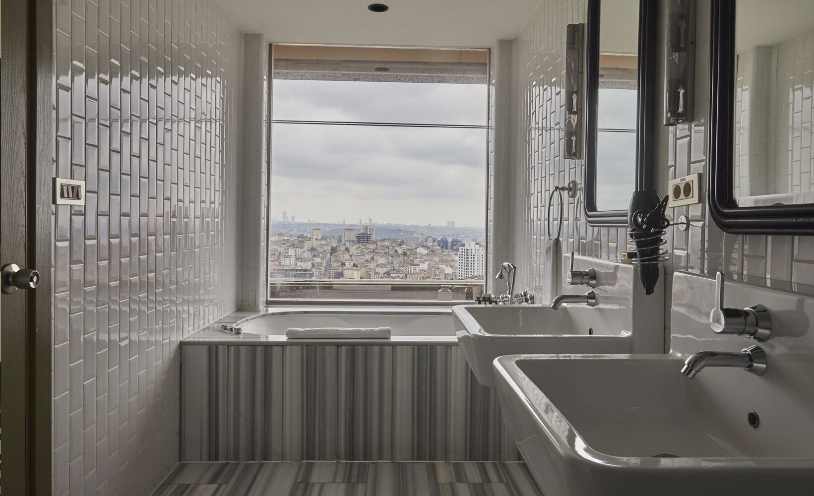 A modern bathroom with two sinks, a bathtub, and a large window offering a city view.