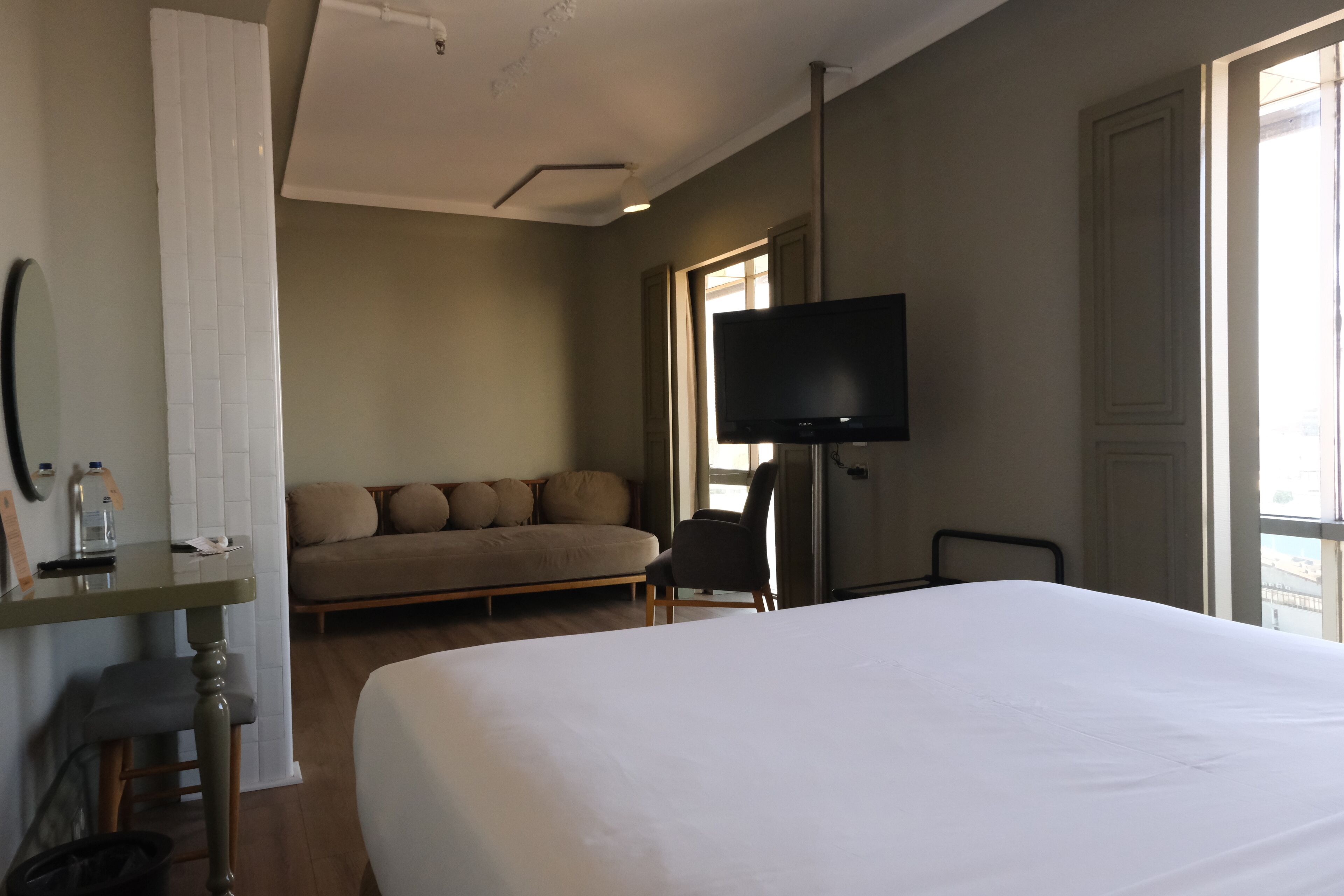 A hotel room with a large bed, a sofa, a television, and a window with natural light.