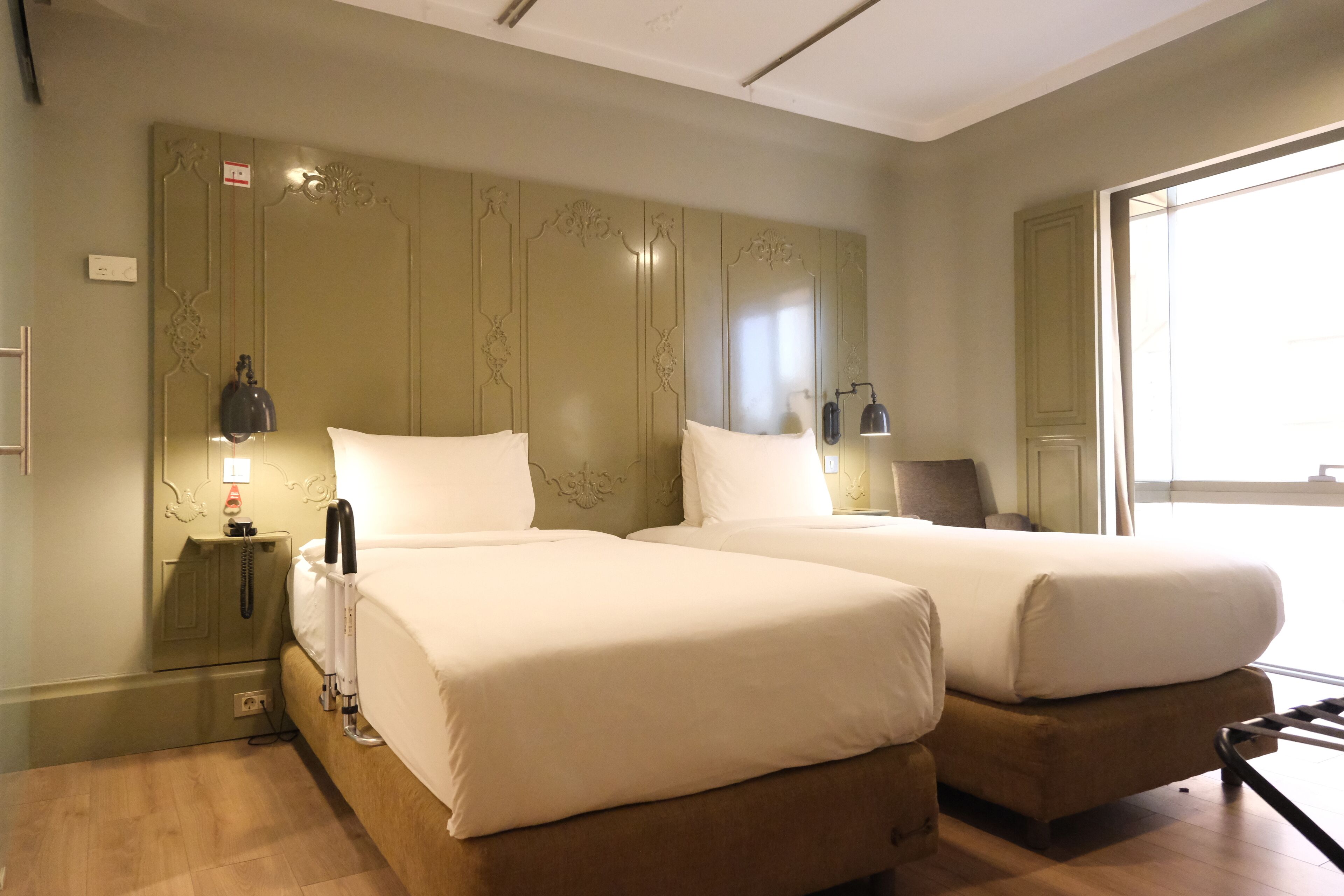 Ein Hotelzimmer mit zwei Betten, einem Nachttisch mit Lampe, einem kleinen Tisch und einem Fenster, das für natürliches Licht sorgt.