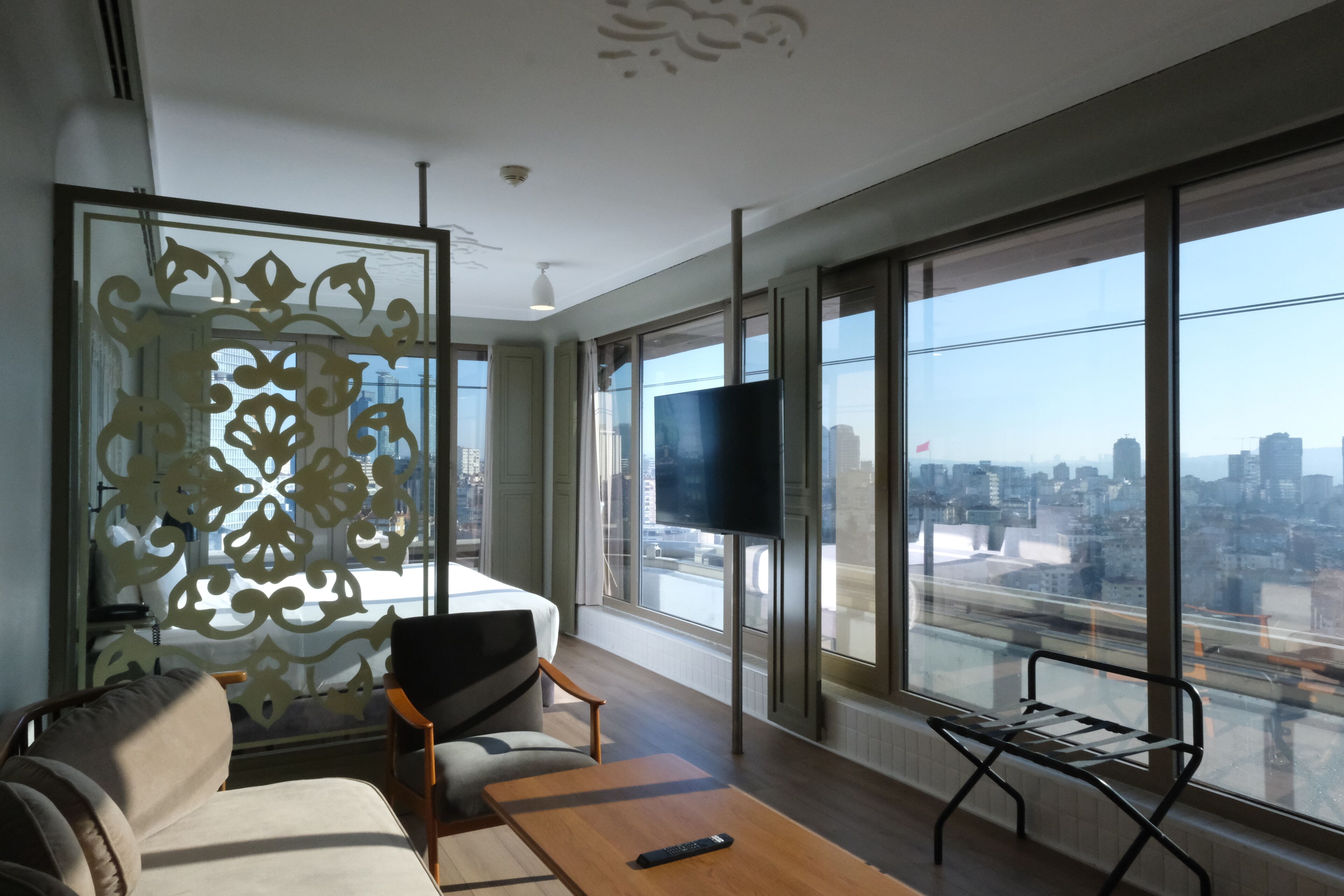 A modern hotel room with a large window, a flat-screen TV, a sofa, a coffee table, and a decorative partition.