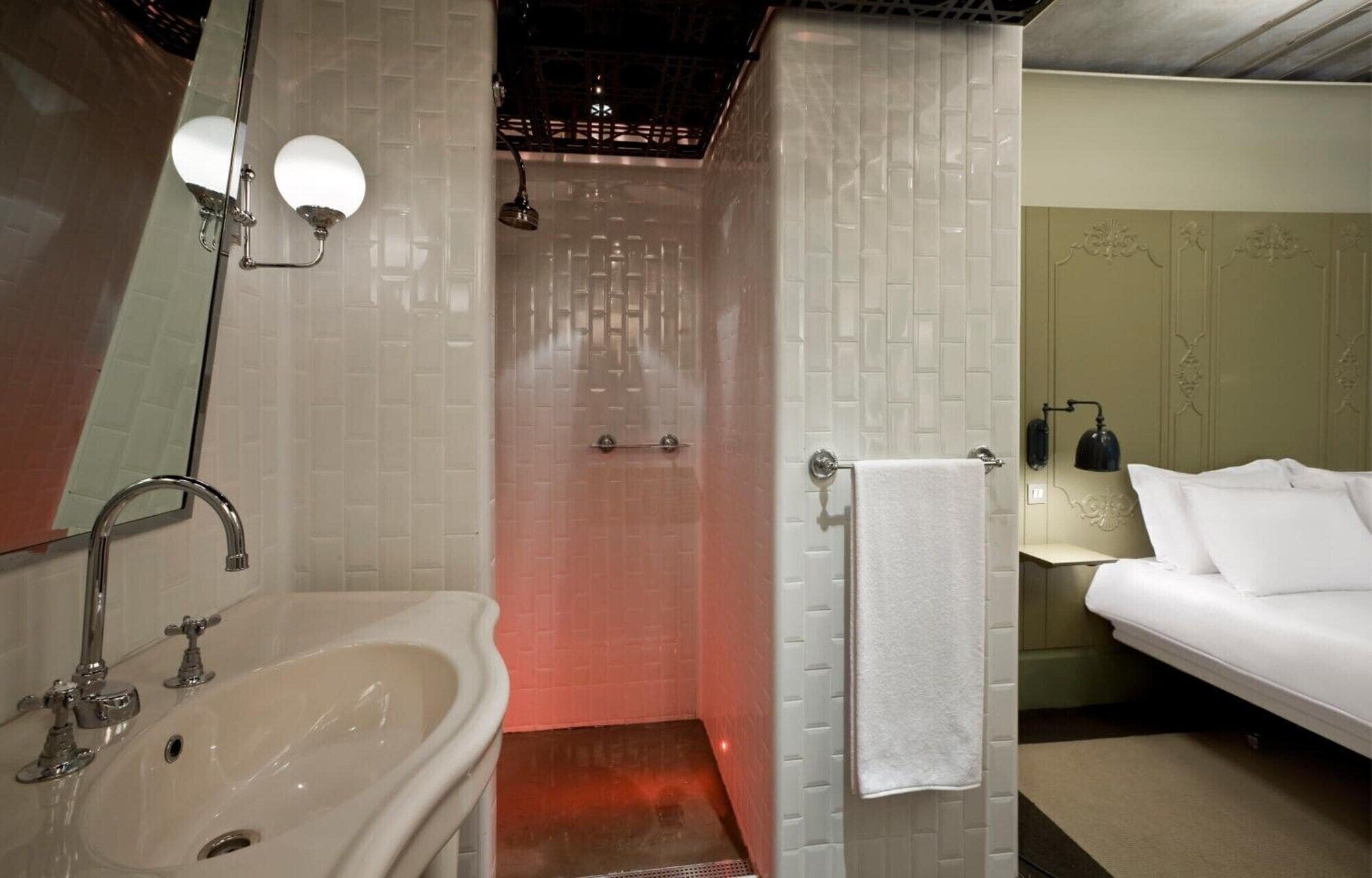 A bathroom with a sink, mirror, shower, and a bed with a towel.
