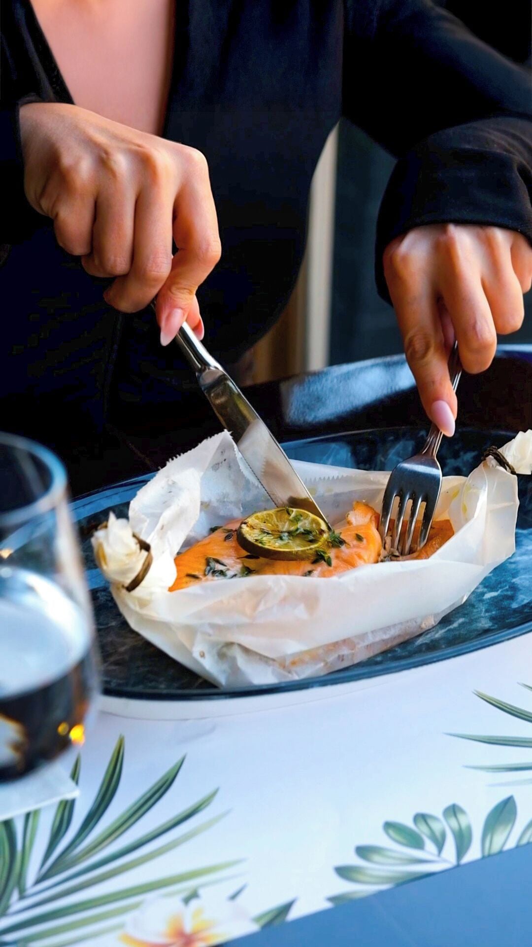 En person spiser en ret med en kniv og gaffel, med et limebåd og krydderurter, serveret på en hvid tallerken med en papirfolie.