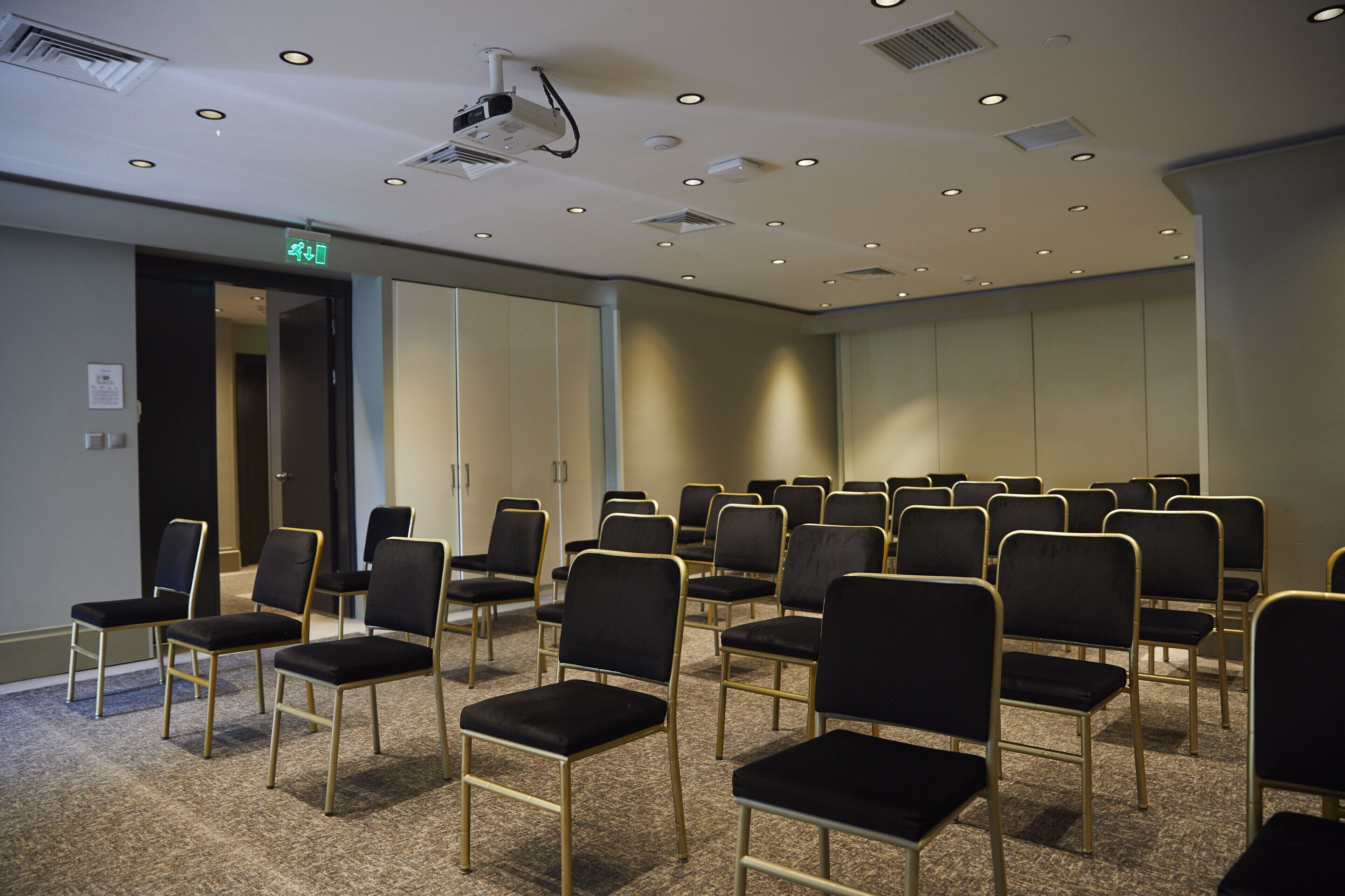 A conference room with rows of black chairs and a projector on the ceiling.