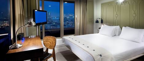 A hotel room with a large bed, a desk with a computer, a chair, a TV, and a city view through the window.
