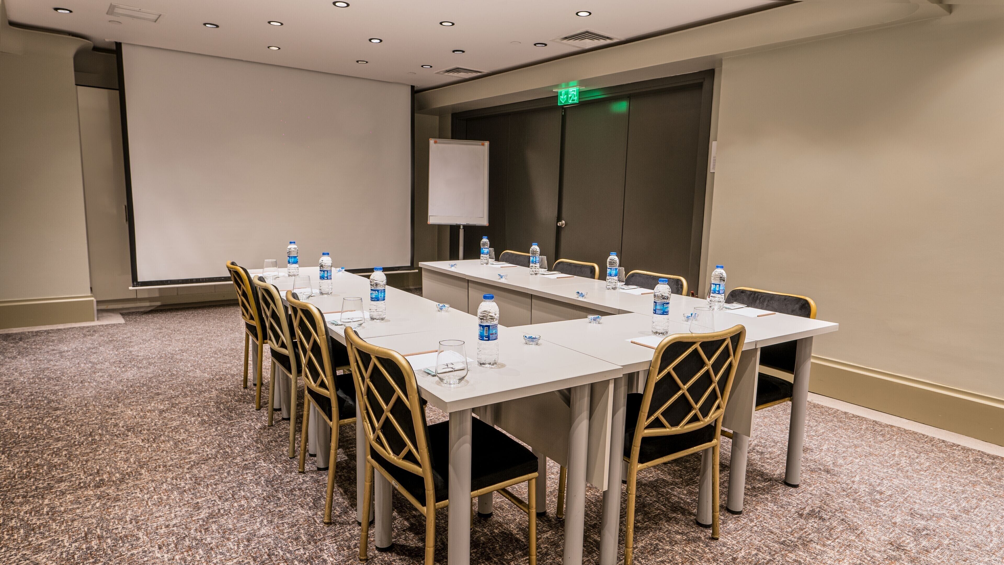 Une salle de conférence avec une longue table, des chaises, des bouteilles d’eau, un écran de projection et une porte portant un panneau indiquant la sortie de secours.