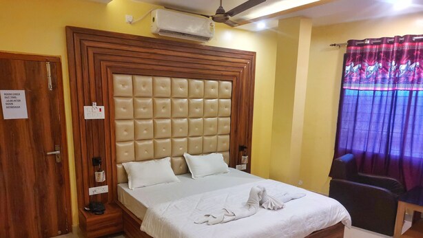 A hotel room with a large bed, wooden headboard, a bedside table, an air conditioning unit, a ceiling fan, and a window with curtains. - Hotel Shobha (Forbesganj)