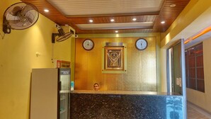 A reception area with a reception desk, wall clocks, a clock with a 'RECEPTION' sign, a wall-mounted fan, and a refrigerator. - Hotel Shobha (Forbesganj)