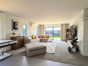 A modern living room with a sectional sofa, a small table, a TV, and a balcony with curtains. - Hideaway in Green Hill Park (Hamilton)