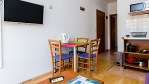 Une petite pièce bien éclairée, avec une table à manger en bois, quatre chaises, un téléviseur à écran plat fixé au mur, un four à micro-ondes posé sur une étagère et une poubelle.