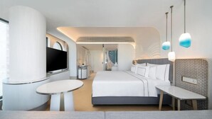 A modern hotel room with a large bed, a TV, a desk, and a seating area.