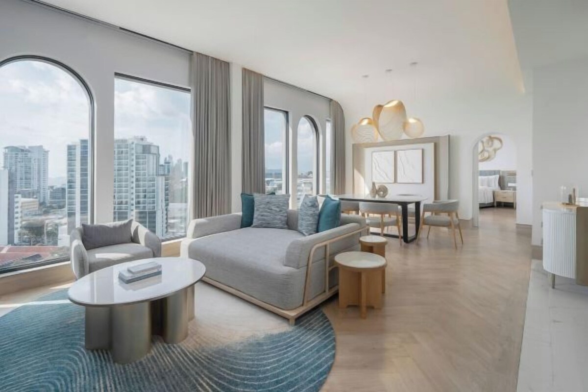 Un salon moderne comprenant un canapé, une table basse et un coin repas, de grandes fenêtres offrant une vue sur la ville, et un style de décoration minimaliste.