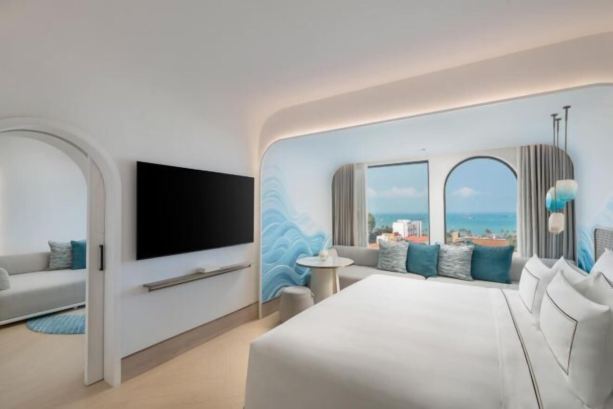 A modern hotel room with a large bed, a flat-screen TV, a sofa, and a view of the sea.