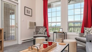 A cozy living room with a grey sofa, a wooden coffee table with mugs, and a view of greenery through large windows.