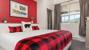 A neatly made bed with a red and black checkered blanket, a white pillow with a black bear design, and a red and black pillow. There is a framed picture on the wall, a small plant on the nightstand, and a window with curtains.