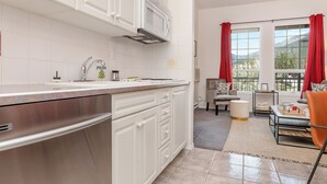 A modern kitchen with white cabinetry, a stainless steel refrigerator, and a microwave. There is a dining area with a table and chairs, and a view of mountains through large windows with red curtains.