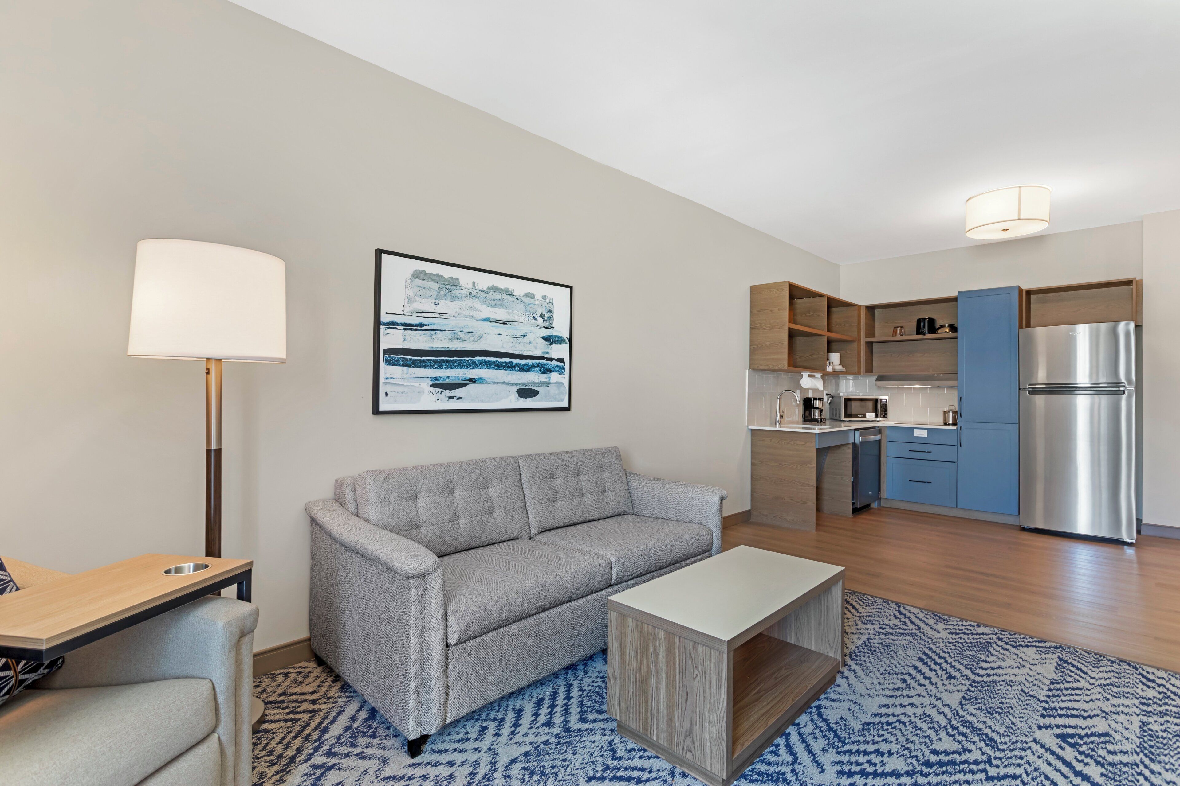 A modern living room with a sofa, a coffee table, and a kitchen area with a refrigerator, microwave, and cabinets.