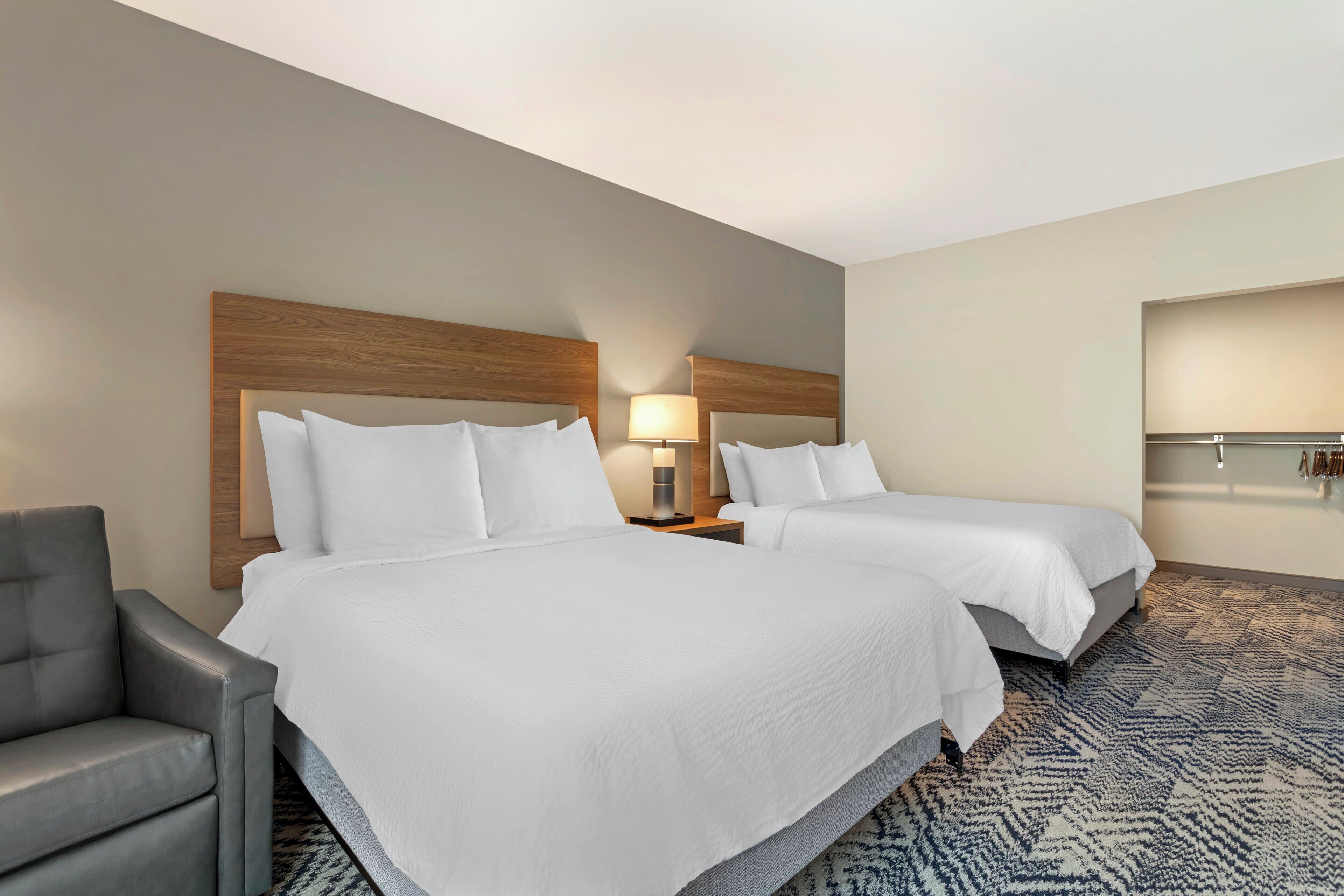 Quarto de hotel com duas camas, uma poltrona cinza, mesa de cabeceira com abajur e tapete estampado.
