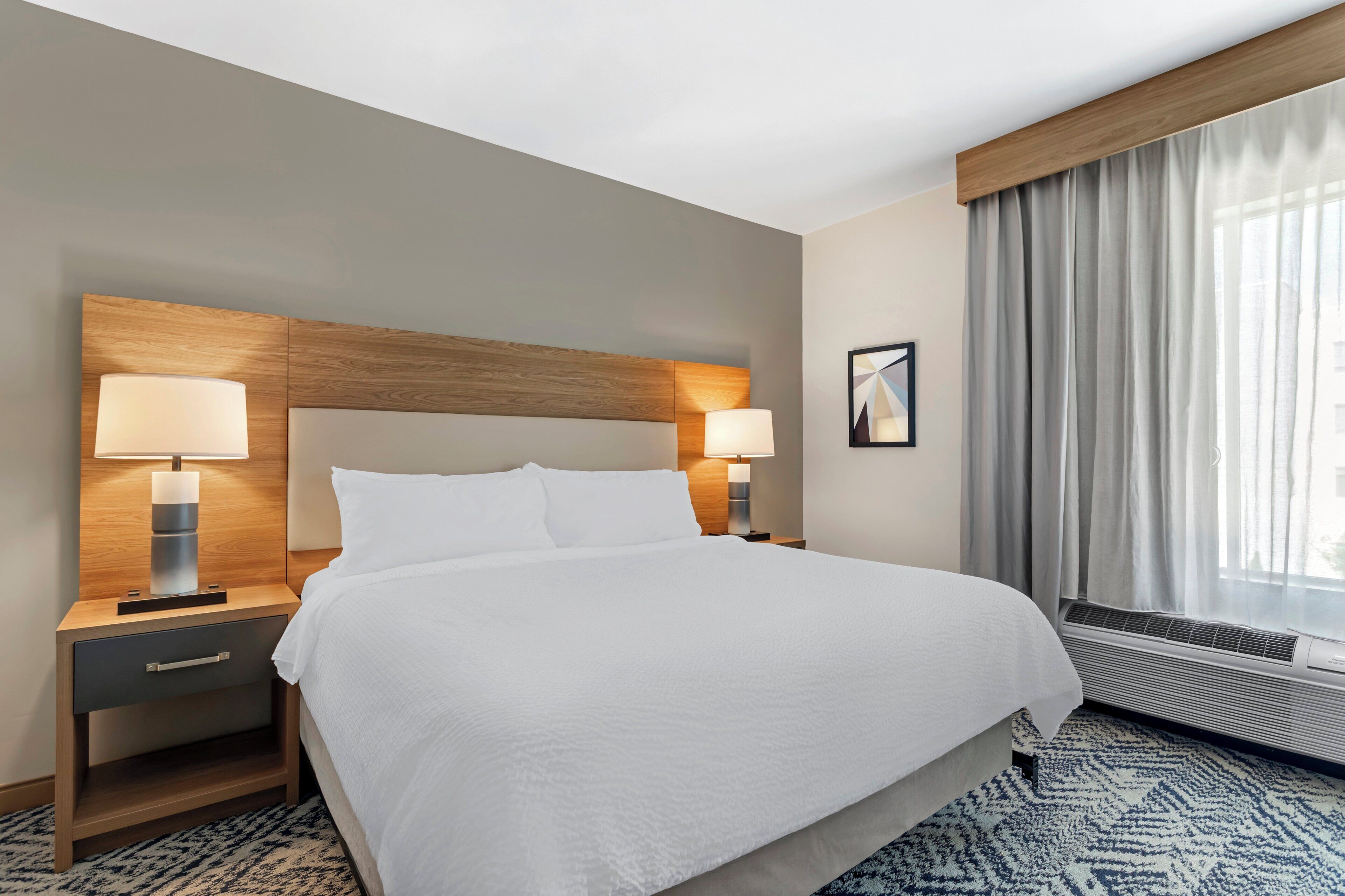 Una habitación de hotel con una cama grande, dos lámparas de noche, una mesita de noche, un cuadro enmarcado en la pared y una ventana con cortinas.
