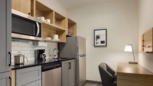 A compact kitchen with a microwave, coffee maker, and a refrigerator. There is a wooden shelf above the sink with cups and glasses. A framed picture hangs on the wall, and a desk with a lamp is visible. - Candlewood Suites Morristown West by IHG (Morristown)