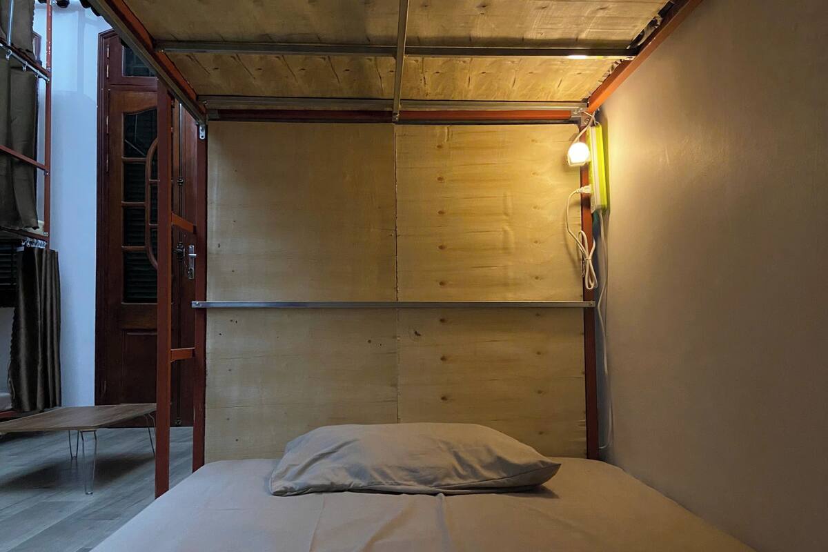 Una habitación pequeña con cama individual, cabecera de madera, lámpara de noche y ventana con cortinas.