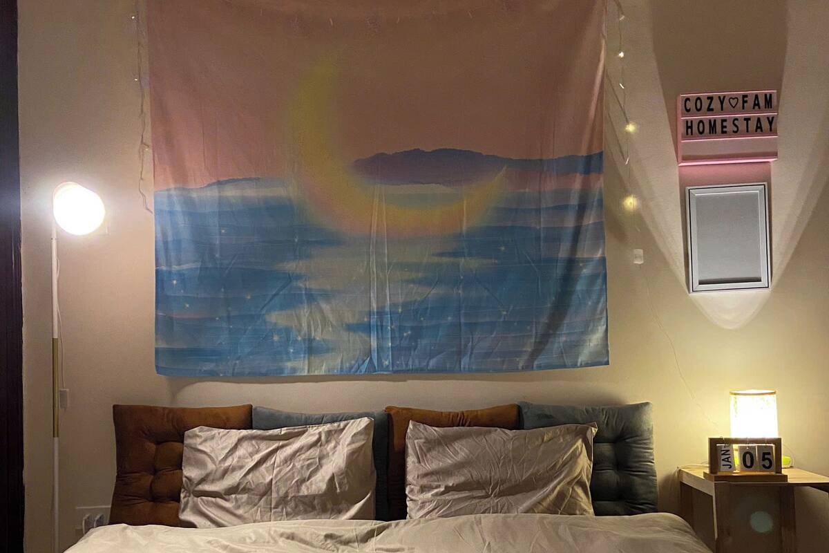 Una cama con sábanas blancas, un cabecero con varios almohadones, un tapiz en la pared, una mesita de noche con una lámpara y un letrero que dice 'COZY HOMESTAY'.
