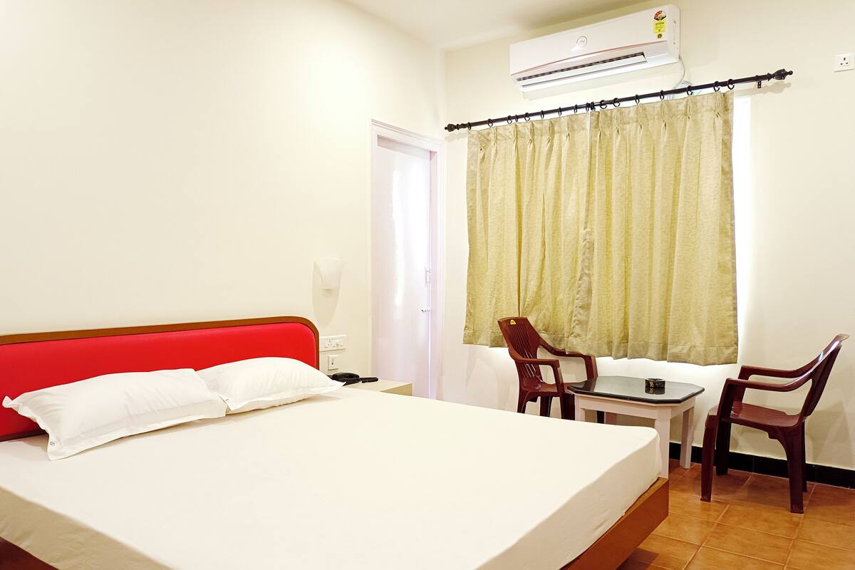 A hotel room with a bed, a table, two chairs, a window with curtains, an air conditioning unit, and a door.