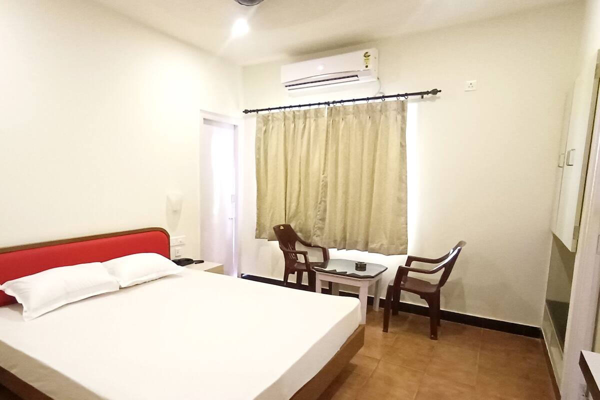A hotel room with a bed, a desk, two chairs, an air conditioning unit, and a window with curtains.