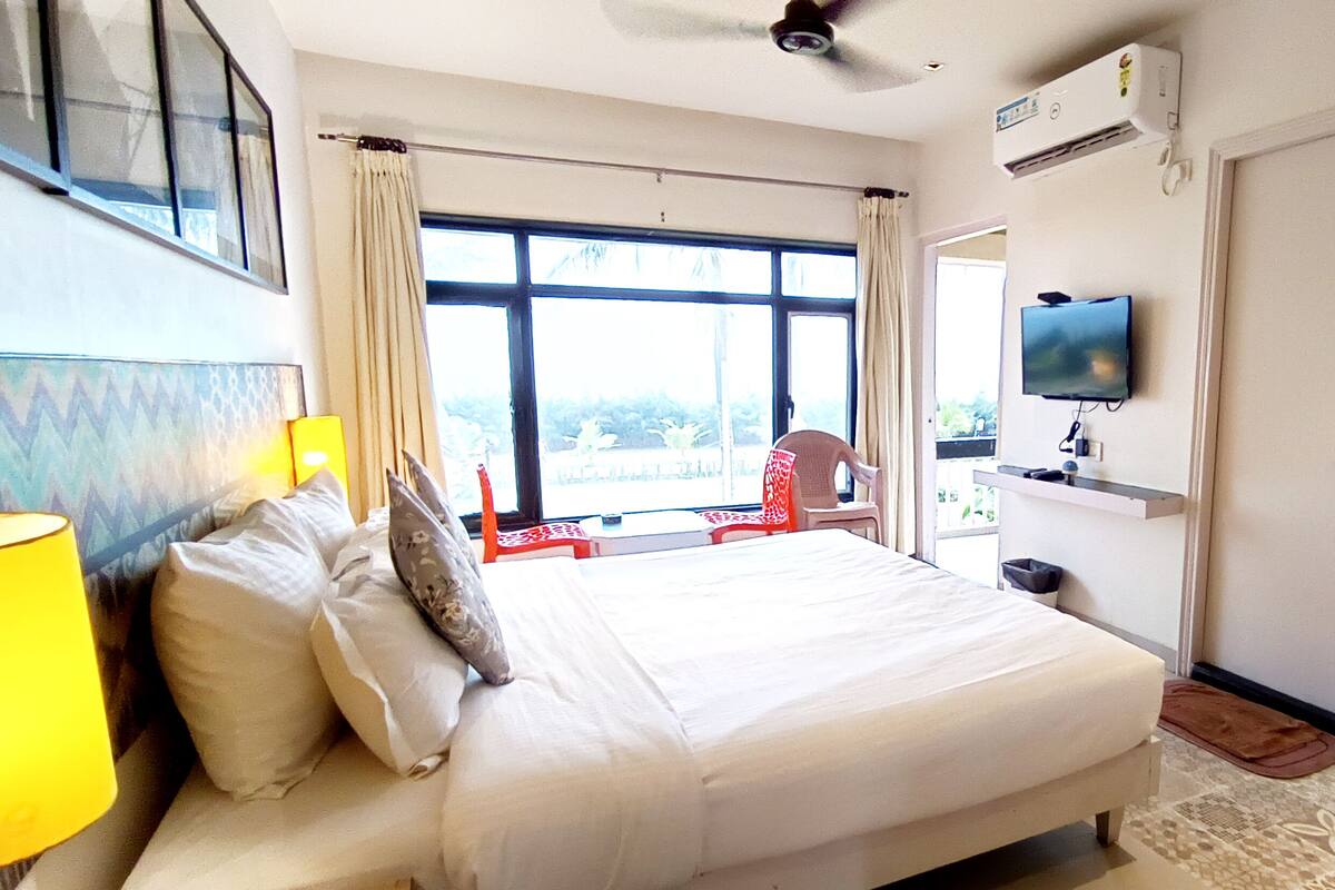 A hotel room with a bed, a TV, a window with curtains, a ceiling fan, and a view of the outdoors.