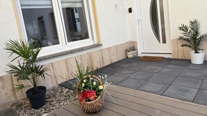 A house entrance with a sliding glass door, a potted plant, a wicker basket with flowers, and a small decorative birdhouse. - Melanie vacation apartment near Bad Ems thermal baths (Bad Ems)